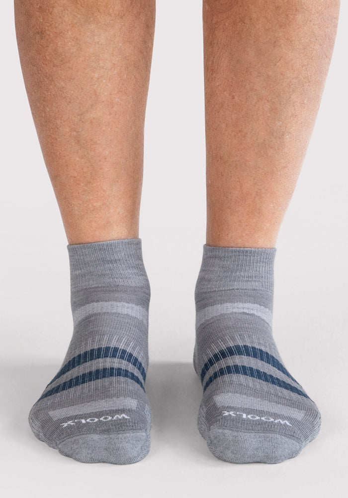 A pair of feet in Woolx Mens Rambler Ankle Sock - Grey with dark blue stripes stands on a light gray surface. These socks have "WELLNESS" on the toes and provide excellent moisture management for all-day comfort.