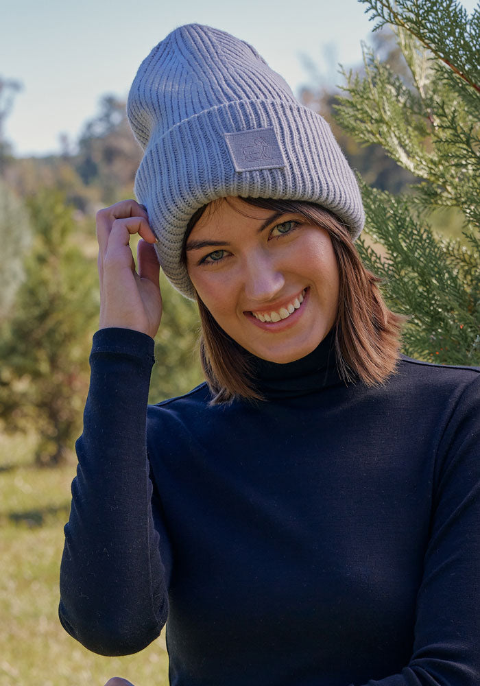 A woman smiles outdoors on a sunny day, wearing the Woolx Ria Chunky Beanie in Morning Fog and a black turtleneck, standing beside green trees.