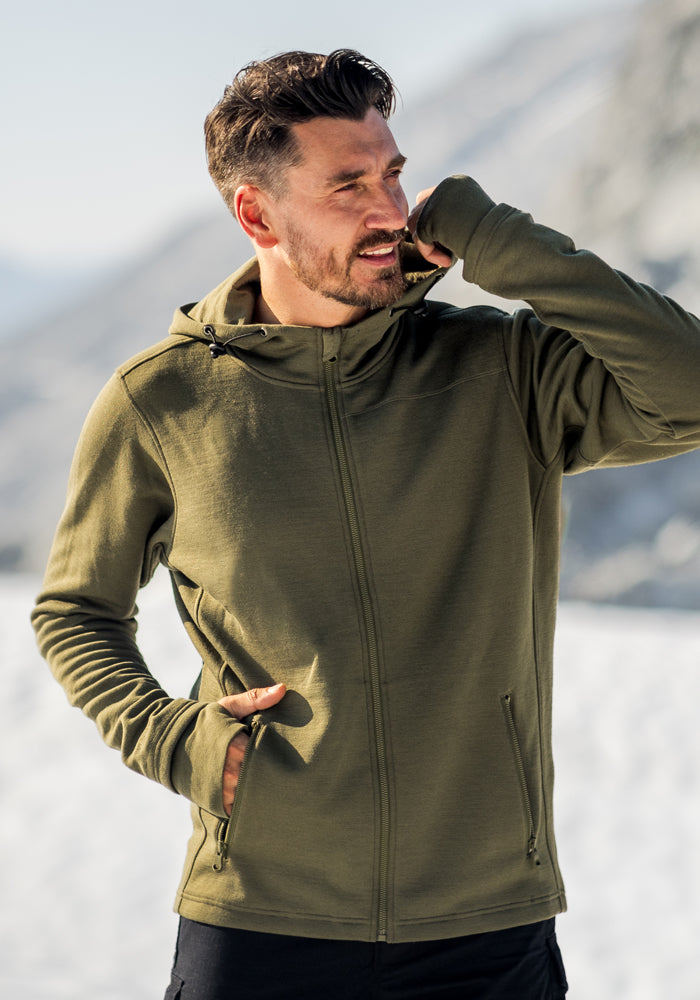 A man wearing the Woolx Grizzly Sweatshirt in Winter Moss stands outdoors in a snowy mountain landscape, one hand in his pocket and the other touching his face.