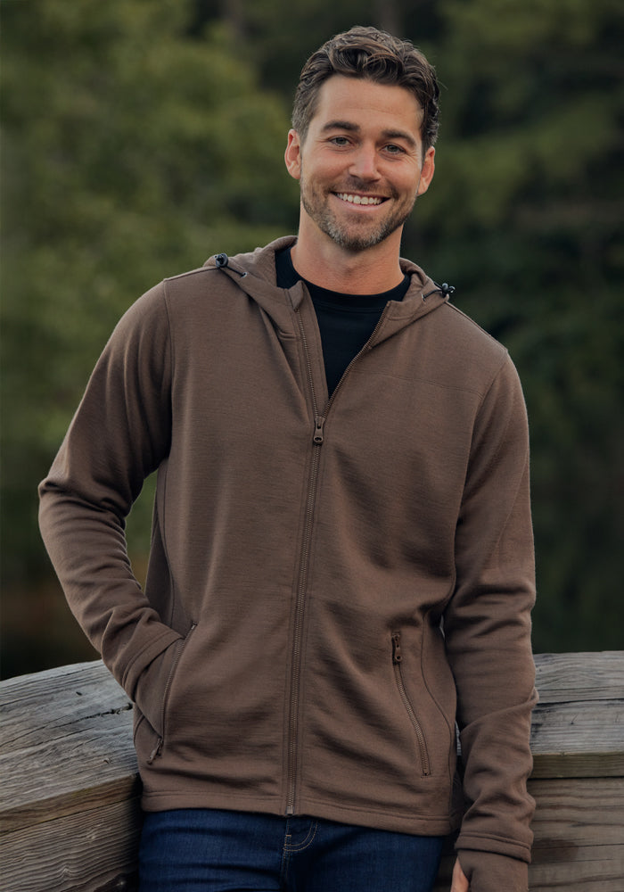 A man with short brown hair and a beard is smiling outdoors by a wooden railing, wearing jeans and the Woolx Grizzly Sweatshirt - Bourbon Oak, with a blurred green background of trees.