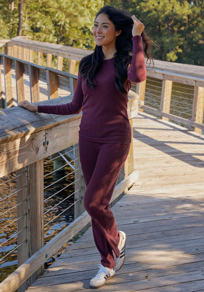 A woman smiles and holds her hair while wearing Woolx's Luca Ribbed Pants in Deep Timber, a burgundy long-sleeve top, and white sneakers as she stands on a wooden boardwalk surrounded by trees.