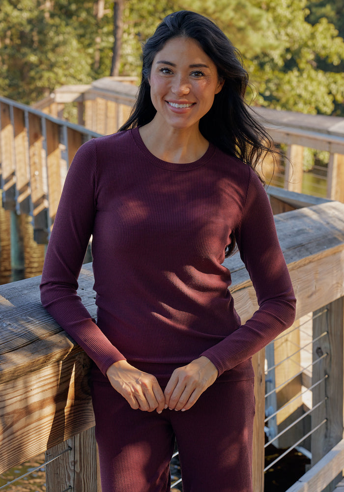 A woman with long dark hair smiles while leaning against a wooden railing outdoors, wearing the Woolx Hadley Ribbed Crew in Majestic Purple, an odor-resistant, moisture-wicking shirt. Sunlight filters through trees in the background.