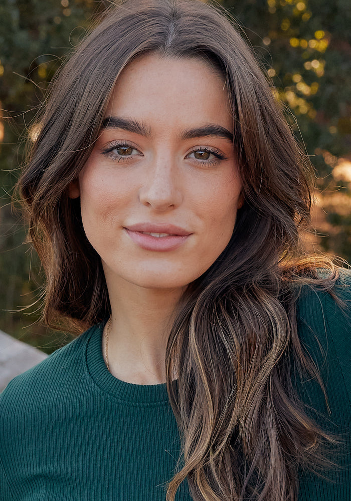 A young woman with long brown hair and light skin wears the Woolx Hadley Ribbed Crew in Deep Timber, standing outdoors and smiling gently at the camera amid sunlight and greenery.