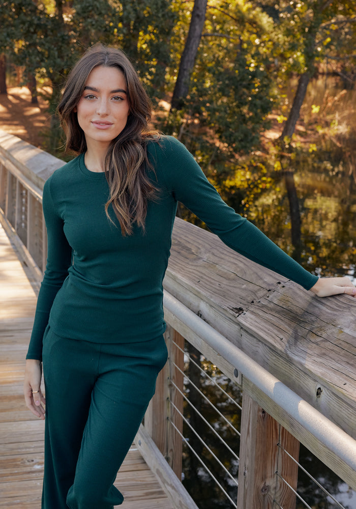 A woman with long brown hair wears the Woolx Hadley Ribbed Crew in Deep Timber, standing on a wooden bridge and leaning on the railing with trees and water behind her.