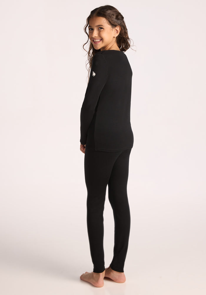 A young girl with long curly hair smiles at the camera, standing barefoot and turned slightly to the side, wearing Woolx Kids Explorer Base Layer Bottoms in black against a plain light background.