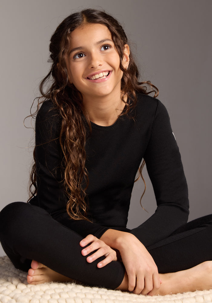 A young girl with long, wavy brown hair smiles while sitting on a cream blanket, wearing the Woolx Kids Explorer Base Layer Top in black, crafted from Australian Merino wool, against a plain neutral background.