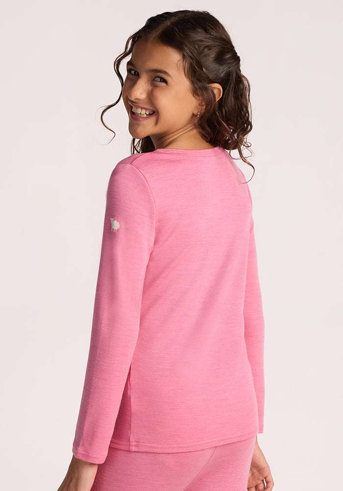A young girl with wavy brown hair smiles over her shoulder, wearing the Woolx Kids Explorer Base Layer Top in Pinky Promise, standing against a plain light background.