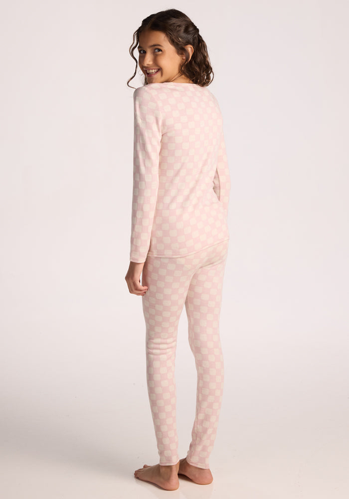 A young girl with wavy brown hair stands barefoot, smiling over her shoulder. She wears Woolx Kids Explorer Base Layer Bottoms in Hopscotch Pink, viewed from the back against a plain white background.