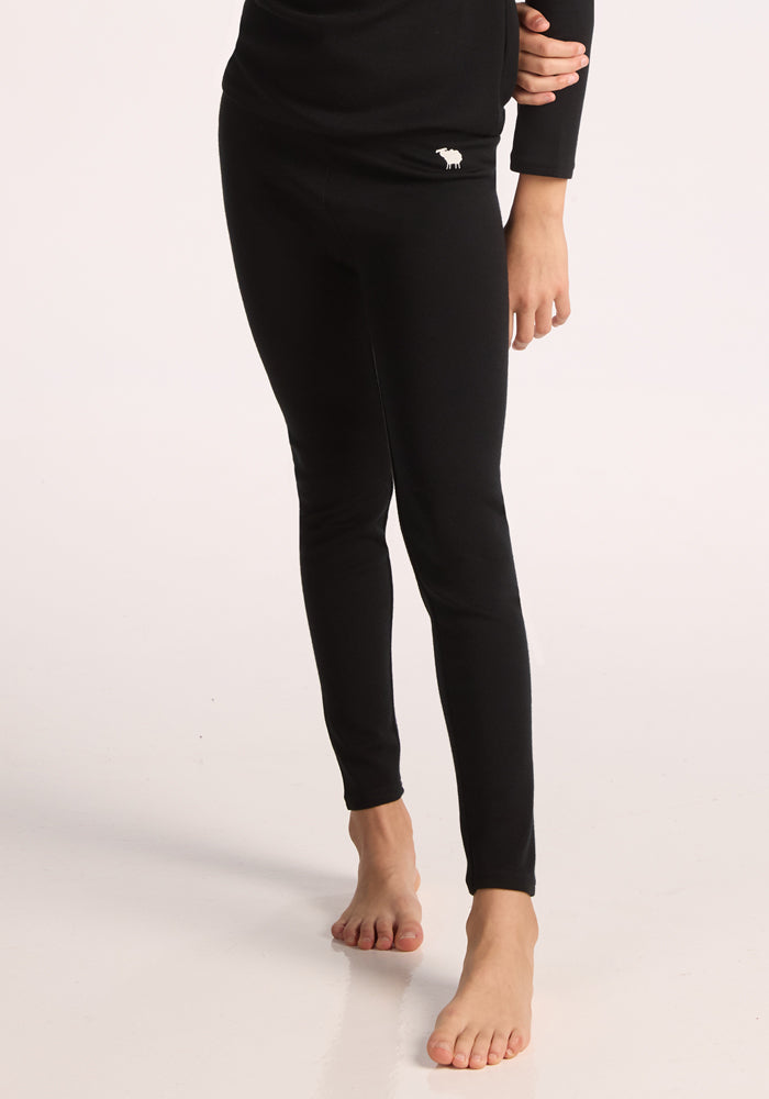 A person stands barefoot on a light surface, wearing Woolx Kids Explorer Base Layer Bottoms in black. Only the lower half is visible, showing a small white animal logo on the upper thigh.