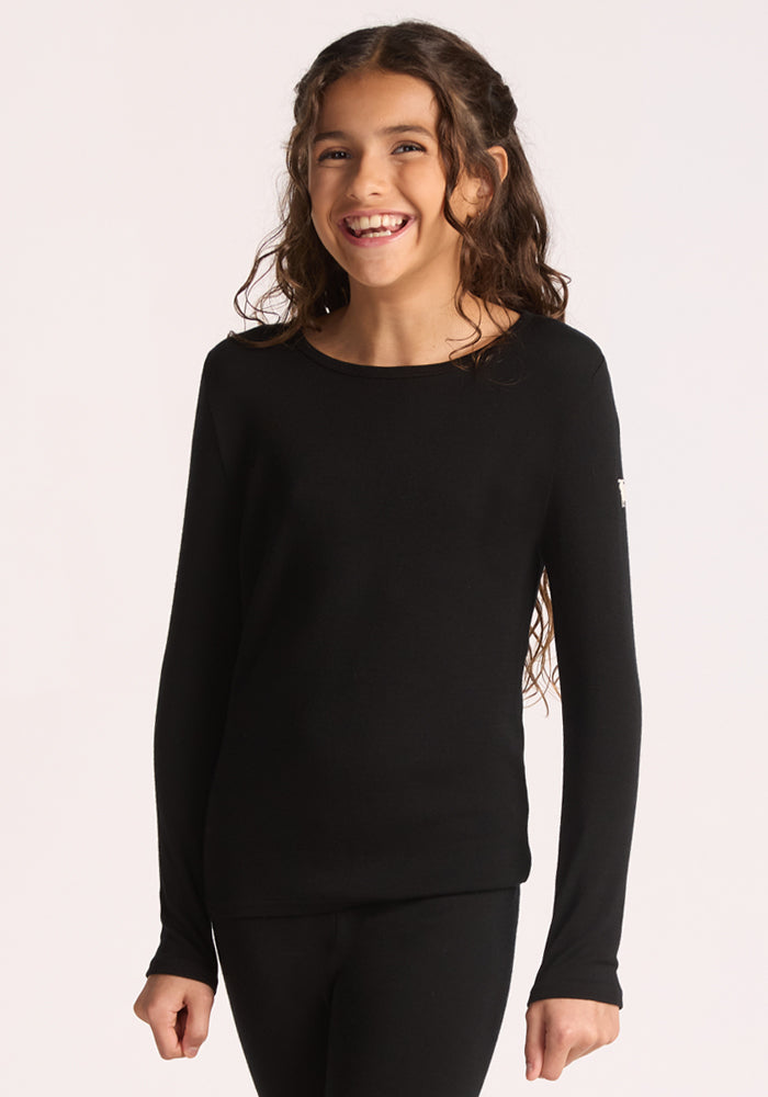 A smiling young girl with long, wavy brown hair wears the Woolx Kids Explorer Base Layer Top in black, made from moisture-wicking Australian Merino wool, standing against a plain light background. 