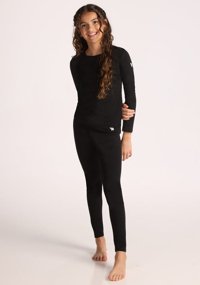 A young girl with long curly hair stands barefoot, smiling, and wearing Woolx Kids Explorer Base Layer Bottoms in black against a plain light background. 