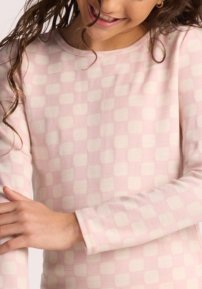 A child smiles in the Woolx Kids Explorer Base Layer Top - Hopscotch Pink. Only the lower face and torso are visible, with long curly hair draped over one shoulder.