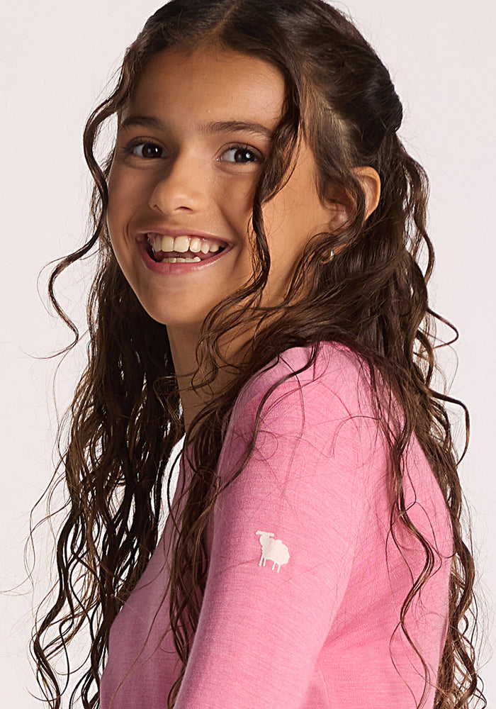 A smiling girl with long, wavy brown hair wears the Woolx Kids Explorer Base Layer Top in Pinky Promise, featuring a small white sheep emblem on the sleeve. She glances over her shoulder against a neutral background.