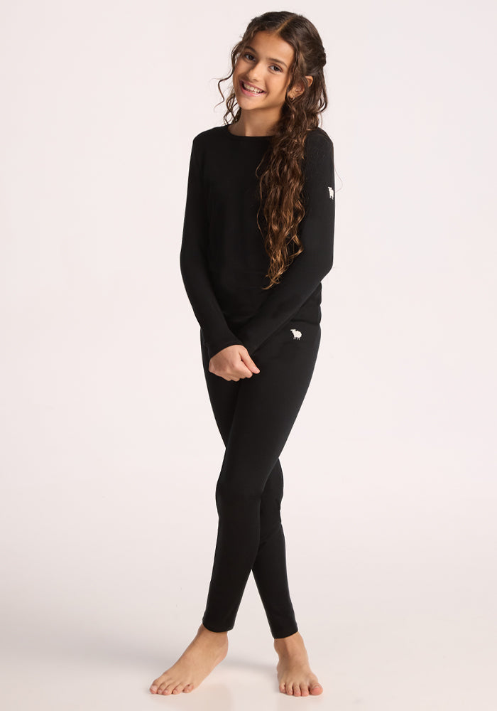 A smiling girl with long curly hair stands barefoot against a light background, wearing the Woolx Kids Explorer Base Layer Top in black, made from moisture-wicking Australian Merino wool.
