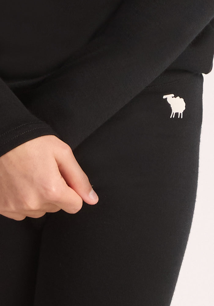 A child wears Woolx Kids Explorer Base Layer Bottoms in black, featuring a small white sheep graphic on the upper thigh as they rest their left hand on their leg.