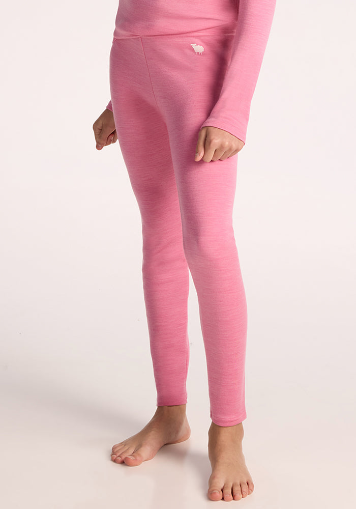A child models Woolx Kids Explorer Base Layer Bottoms in Pinky Promise, barefoot against a plain light background. The warm merino wool leggings feature a small white animal logo near the hip. 