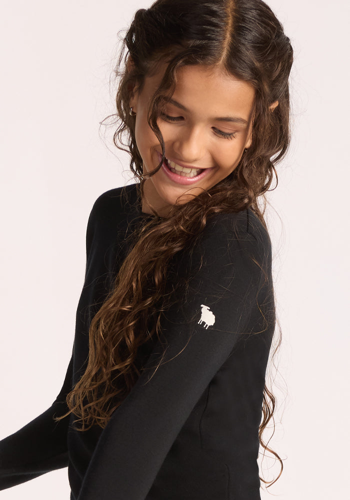 A smiling young girl with long curly hair wears the Woolx Kids Explorer Base Layer Top in black, made from soft Australian Merino wool and featuring a small white sheep logo on the sleeve, standing against a plain light background.