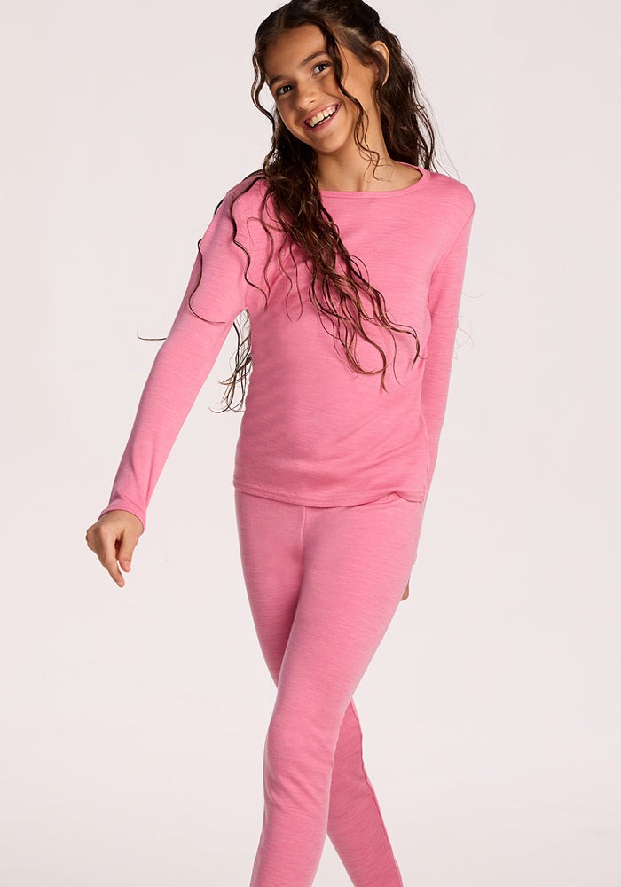 A smiling girl with long brown hair wears Woolx Kids Explorer Base Layer Bottoms in Pinky Promise and a matching top. She stands against a plain light background, extending one arm as if in motion.