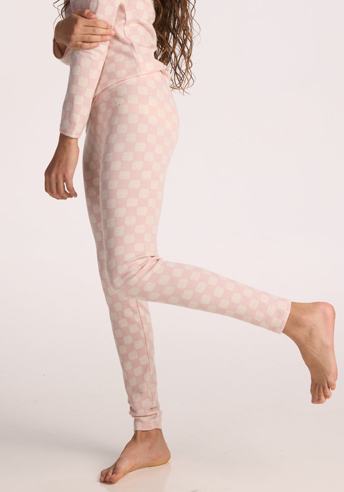 A child models Woolx Kids Explorer Base Layer Bottoms in Hopscotch Pink, standing on one leg in light pink and white checkered bottoms. Only the lower body and part of an arm are shown against a plain background.