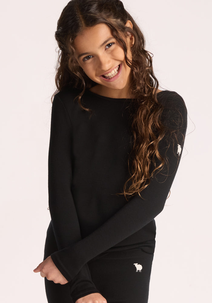 A smiling young girl with long, wavy brown hair wears the Woolx Kids Explorer Base Layer Top in black, made from soft Australian Merino wool and featuring a small white sheep logo, standing against a plain light background.