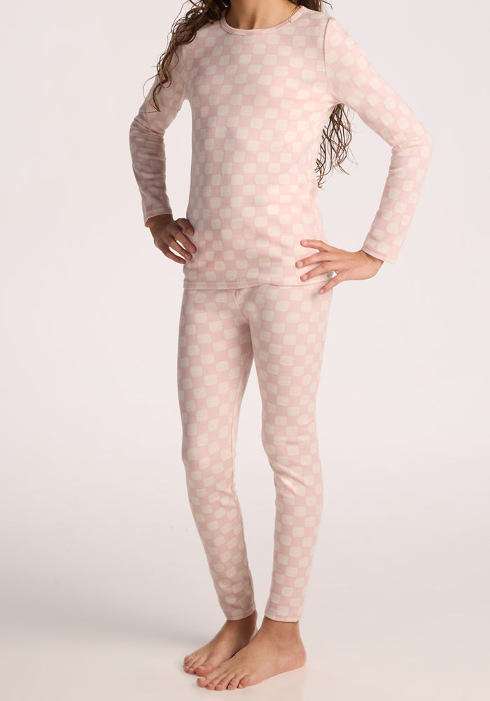 A child stands barefoot on a plain light background, hands on hips, wearing Woolx Kids Explorer Base Layer Bottoms in Hopscotch Pink paired with a matching pink and white checkered long-sleeve top.