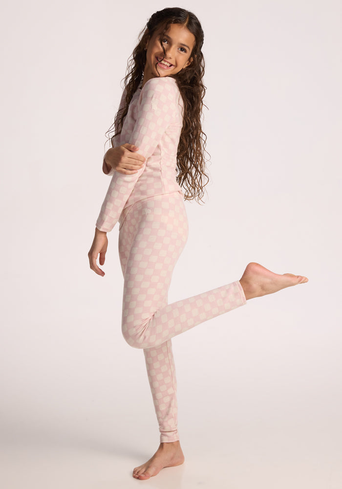 A girl with long curly hair smiles and poses on one leg, wearing the Woolx Kids Explorer Base Layer Top in Hopscotch Pink. The light pink, long-sleeve top features moisture-wicking fabric and a subtle checkered pattern against a plain white background.