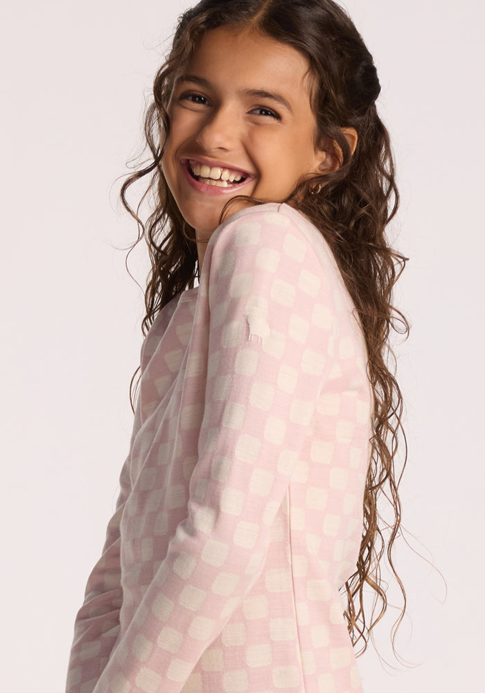 A smiling young girl with long, wavy brown hair wears the Woolx Kids Explorer Base Layer Top in Hopscotch Pink, posing against a plain background—perfect for highlighting the comfort and style of this merino wool essential.