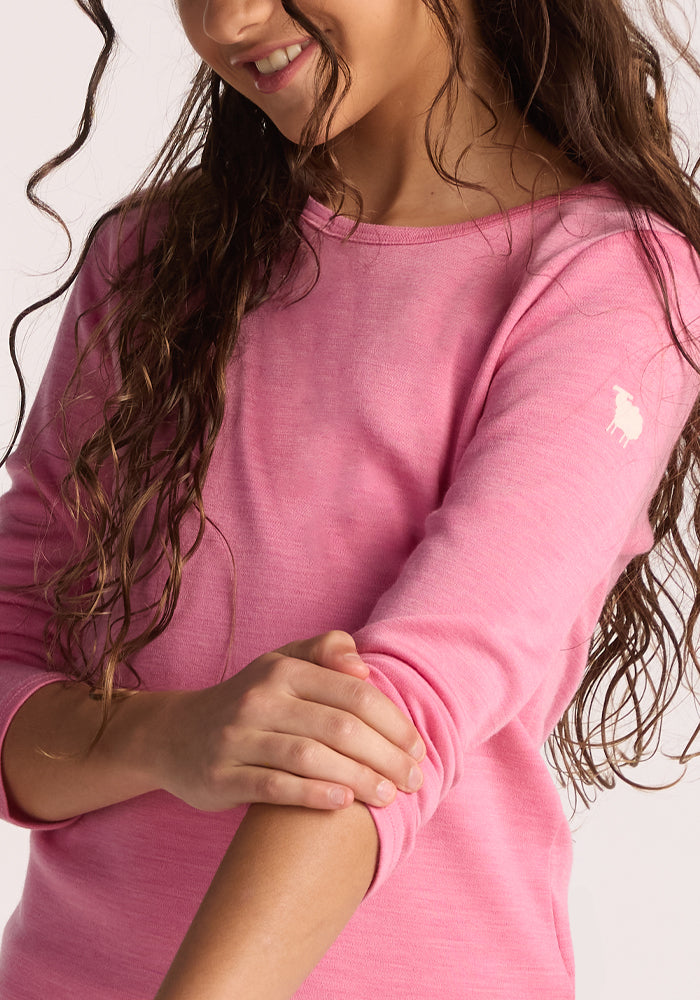 A girl with long, wavy brown hair smiles in the Woolx Kids Explorer Base Layer Top - Pinky Promise, touching her opposite forearm. A white sheep logo on the sleeve shows it's made from soft, moisture-wicking Australian Merino wool.