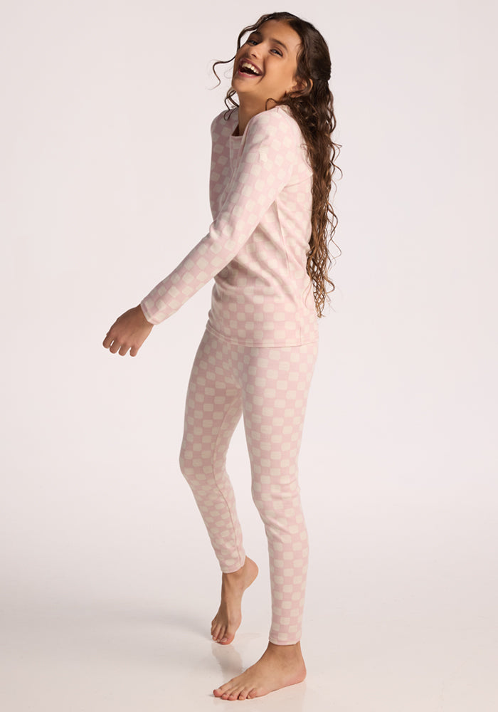 A smiling girl with long curly hair stands barefoot, playfully posing against a plain white background in Woolx Kids Explorer Base Layer Bottoms - Hopscotch Pink.