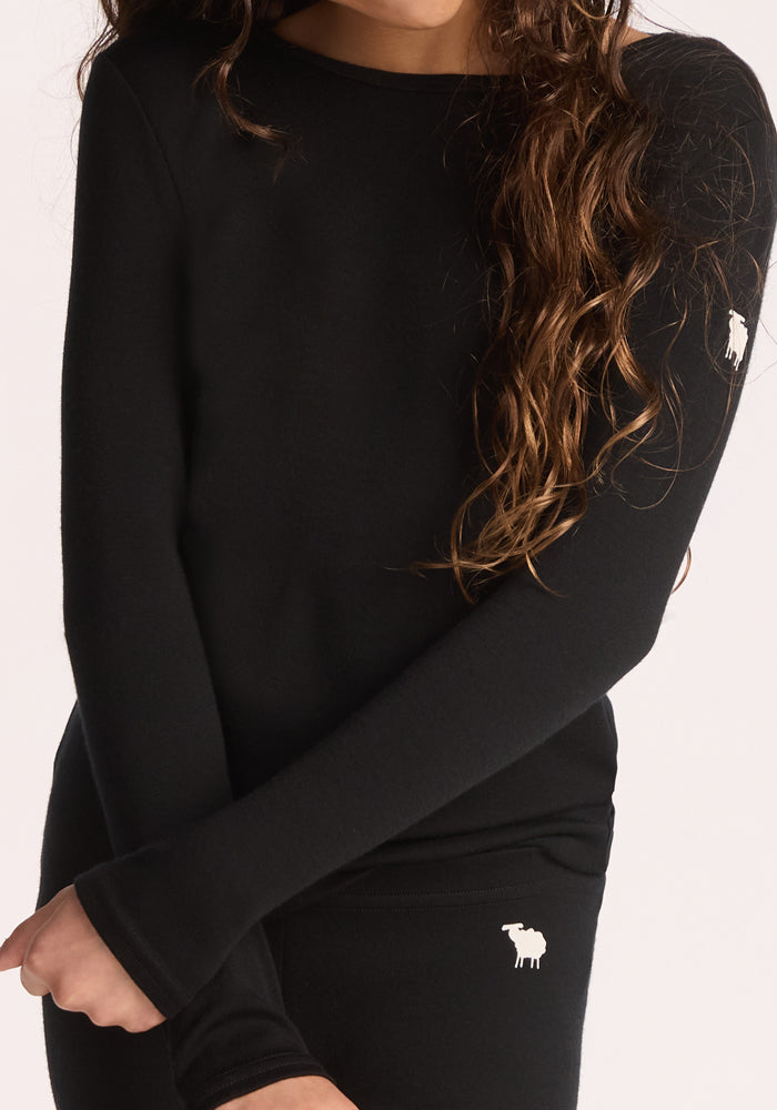A person with long, wavy brown hair wears a fitted black Woolx Kids Explorer Base Layer Top and matching pants, both featuring a small white moose logo. The person's face is not visible.