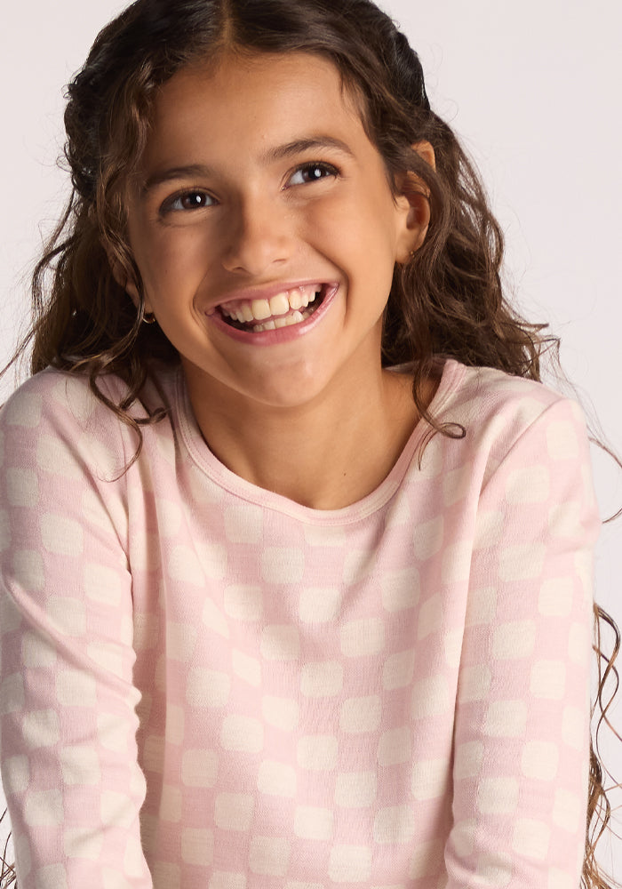 A young girl with long curly brown hair smiles brightly, looking to the side, wearing the Woolx Kids Explorer Base Layer Top in Hopscotch Pink—a cozy merino wool piece providing comfort and warmth against a plain background.