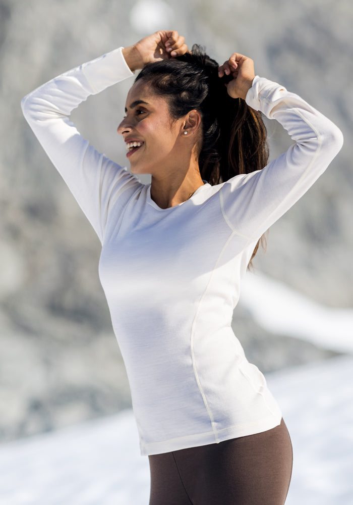 A woman wearing the Woolx Hannah Long Sleeve Top in Buttercream and brown pants stands in snowy mountains, smiling as she ties her hair back.