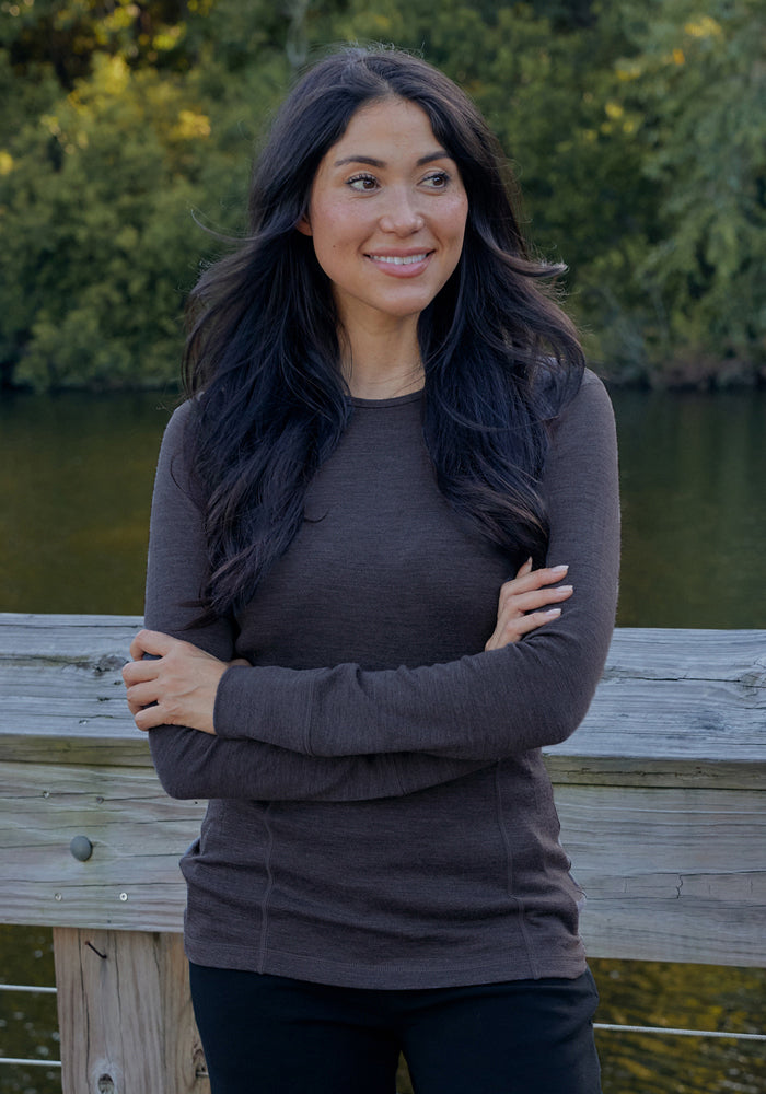 A woman with long dark hair, wearing the Woolx Hannah Long Sleeve Top in Cinder Melange and black pants, stands on a wooden bridge with her arms crossed, smiling to the side. Green trees and water are visible in the background.