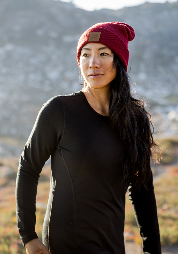A woman with long dark hair wears a Woolx Elsa Long Sleeve top in black and a red beanie, standing outdoors with mountains in the background and looking off to the side.