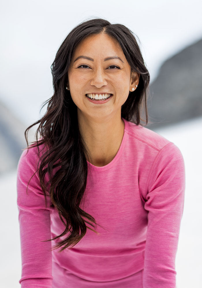 Smiling woman in the Woolx Hannah Long Sleeve Top—Pinky Promise—stands outdoors, long dark hair, blurred background.