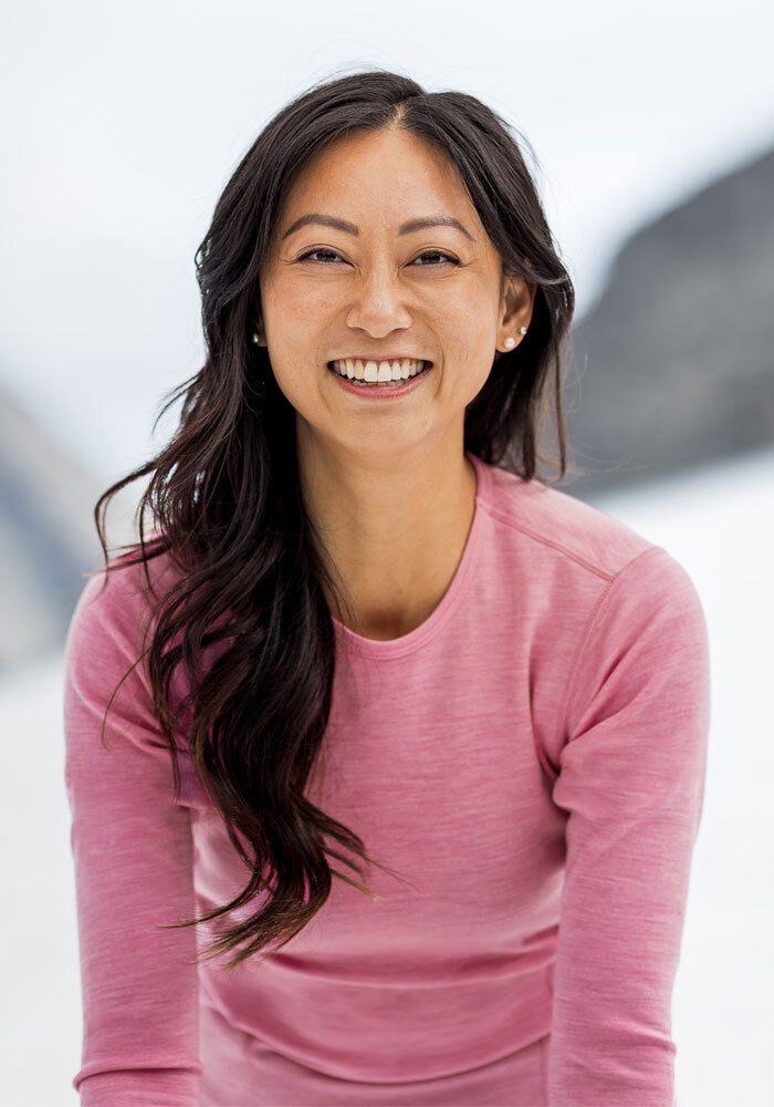 Woman with long dark hair smiles in a Woolx Hannah Long Sleeve Top - Pinky Promise, outdoors with a blurred backdrop.
