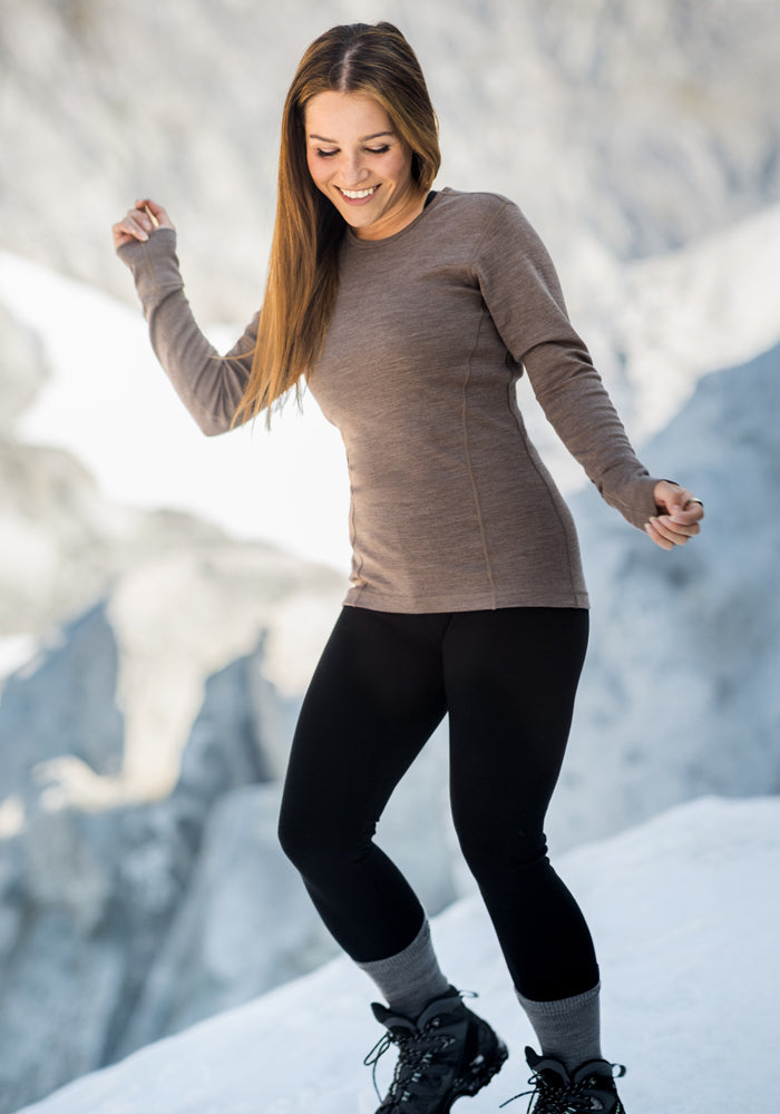 A woman with long brown hair, wearing a brown long-sleeve shirt, Merino wool leggings, gray socks, and Woolx Stella Petite - Black boots, smiles while standing on snow with a blurred rocky background.