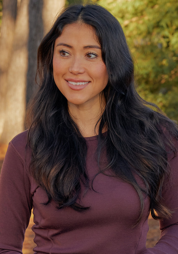 A woman with long, wavy dark hair stands outdoors in the sunlight, smiling and wearing the Woolx Hannah Long Sleeve Top in Chocolate Fig, enjoying its temperature regulating merino wool comfort.