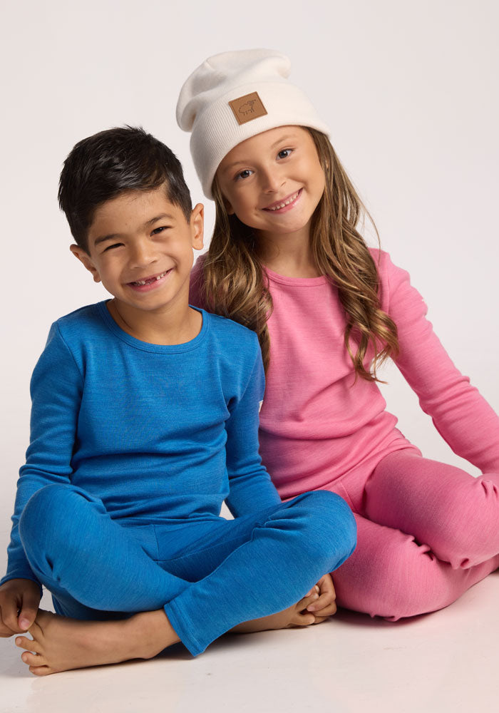 Two children sit cross-legged and smiling. The boy wears a blue outfit, while the girl sports a pink look and the Woolx Kids Baylor Beanie in Whipped Cream. The background is plain white.