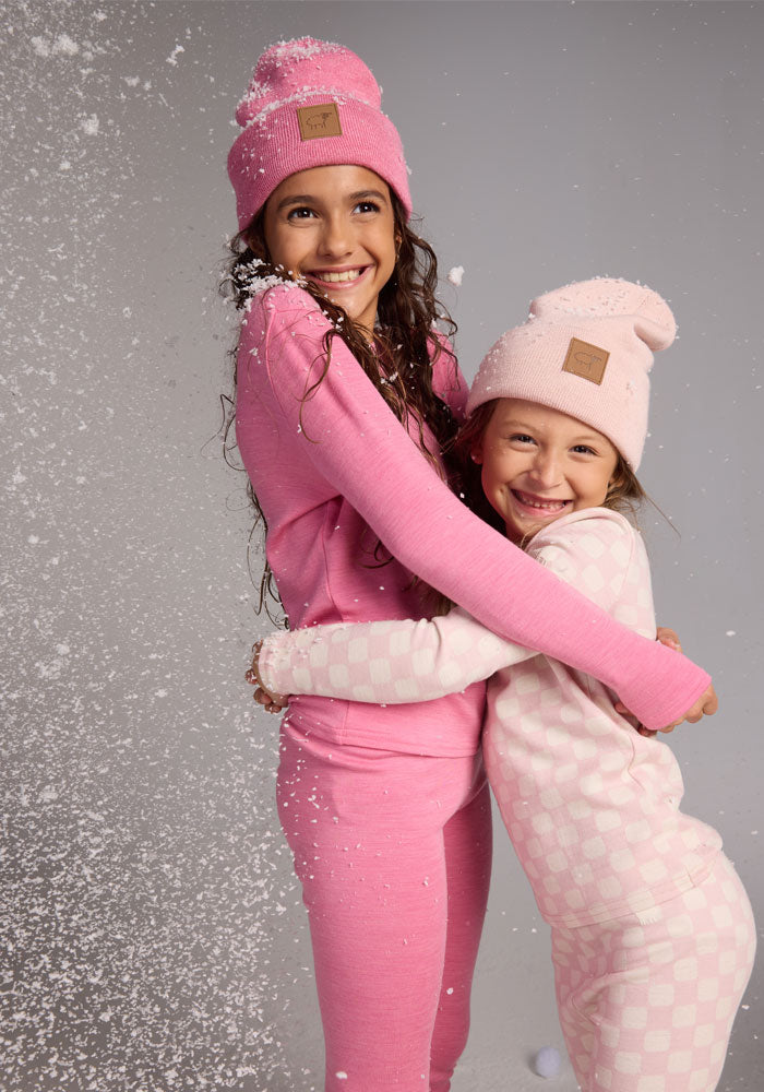 Two smiling girls in pink pajamas and matching Woolx Kids Baylor Beanie - Pinky Promise hats hug as artificial snow falls around them, creating a joyful and playful winter scene.