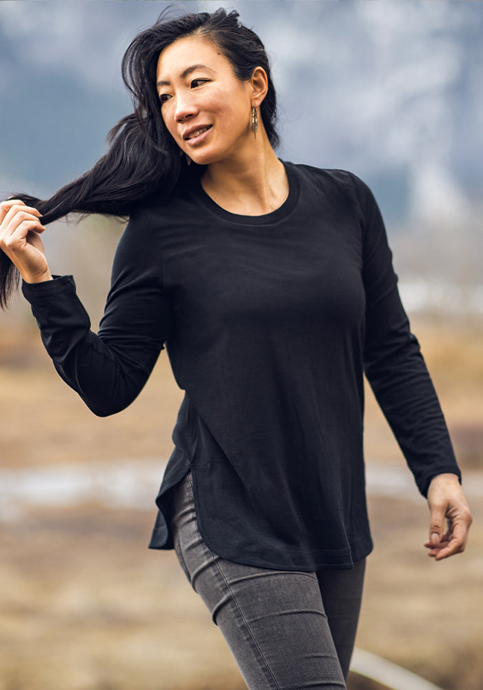 A woman with long dark hair smiles outdoors, wearing the Woolx Hazel Tunic in black and gray jeans. The lightweight, moisture-wicking Merino wool keeps her comfortable as she enjoys the natural surroundings.
