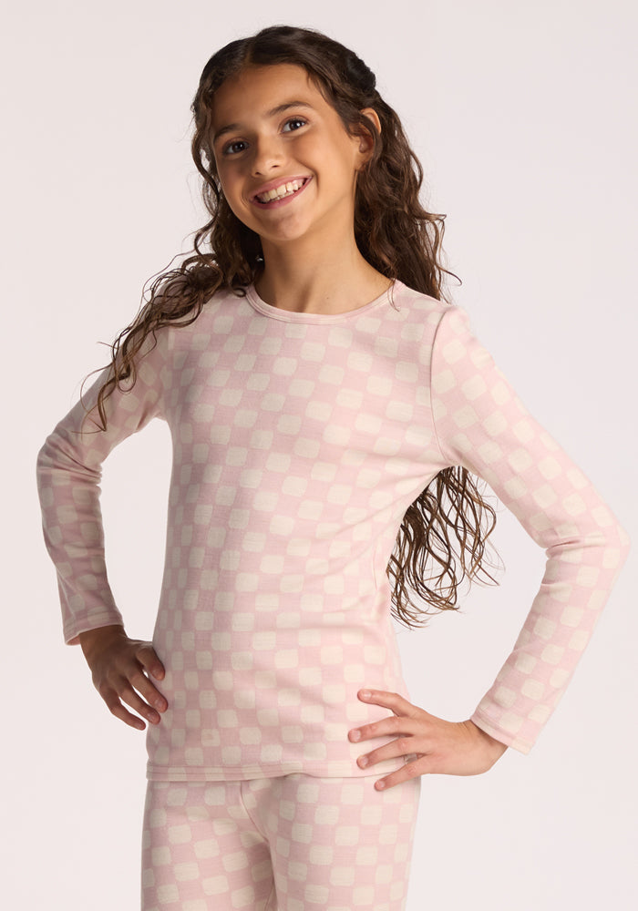 A young girl with long curly hair smiles while posing in the Woolx Kids Explorer Base Layer Top in Hopscotch Pink, featuring long sleeves and matching pants, against a plain light background. 