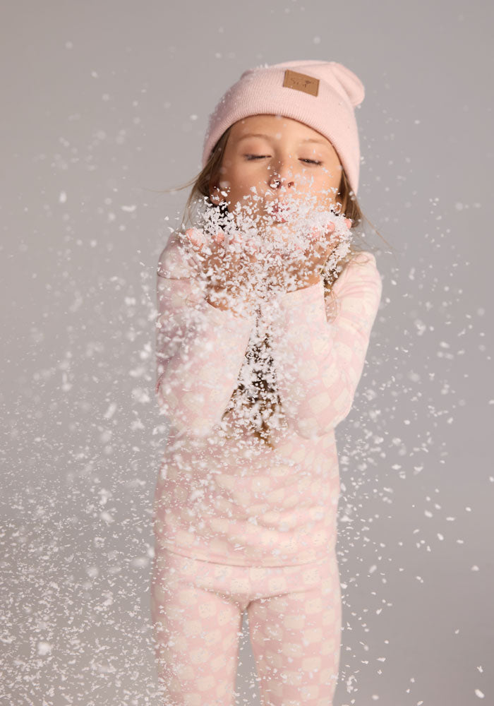 A young girl in a Woolx Kids Explorer Base Layer Top in Hopscotch Pink blows artificial snow towards the camera, creating a playful flurry of white flakes.