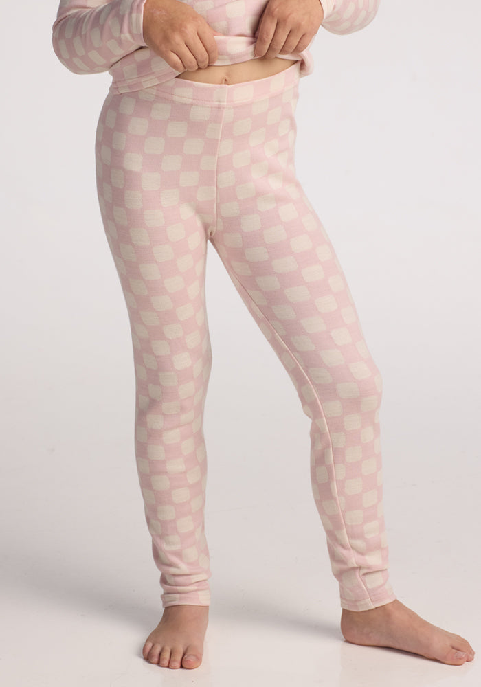 A child stands barefoot on a white background, wearing Woolx Kids Explorer Base Layer Bottoms in Hopscotch Pink checkered print and resting one hand on their waistband. 