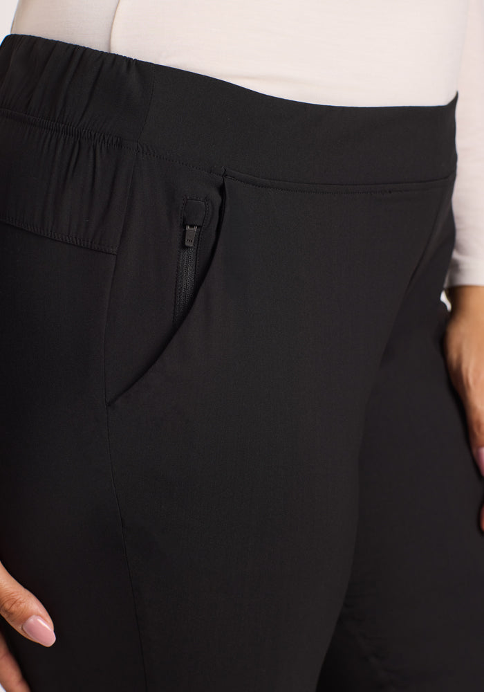 Close-up of a person wearing Woolx Nicole Tall Pants in black, featuring lightweight, breathable fabric, elastic waistband, and zippered pocket. Paired with a light-colored long-sleeve top; the person's hand is near the pocket.