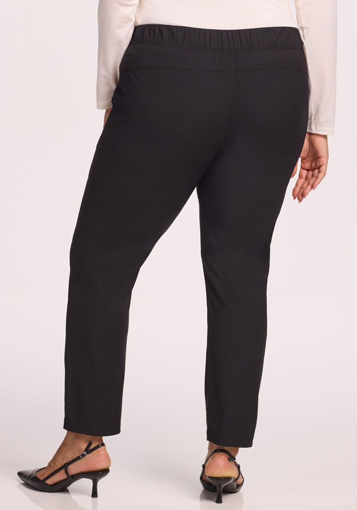 A person is shown from the back wearing Woolx Nicole Tall Pants in black—straight-leg, lightweight, breathable with an elastic waistband—paired with black heeled sandals and a long-sleeved light-colored top.