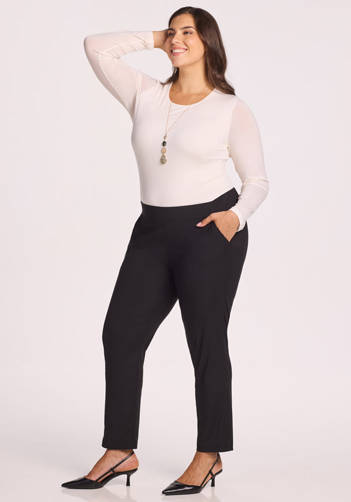 A woman poses confidently with one hand on her head and the other in her pocket, wearing Woolx Nicole Pants in Black, a long-sleeve white top, black heels, and a necklace against a plain light background. 
