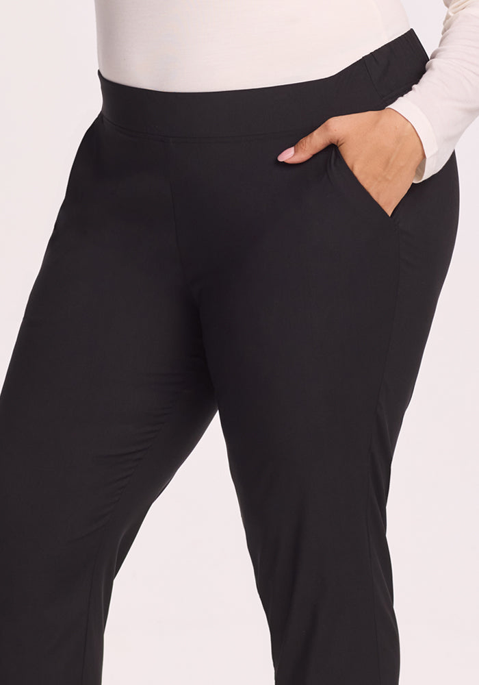 A person models Woolx Nicole Pants in black, paired with a white long-sleeve top, one hand in their pocket, shown from the waist down against a plain background.