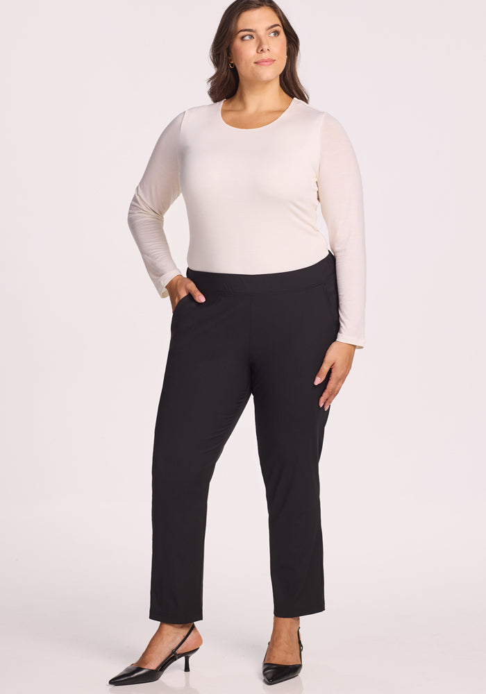 A woman stands against a plain background, wearing Woolx Nicole Pants in black paired with a long-sleeved white top and black heeled shoes. She has one hand in her pocket and looks off to the side with a relaxed expression.