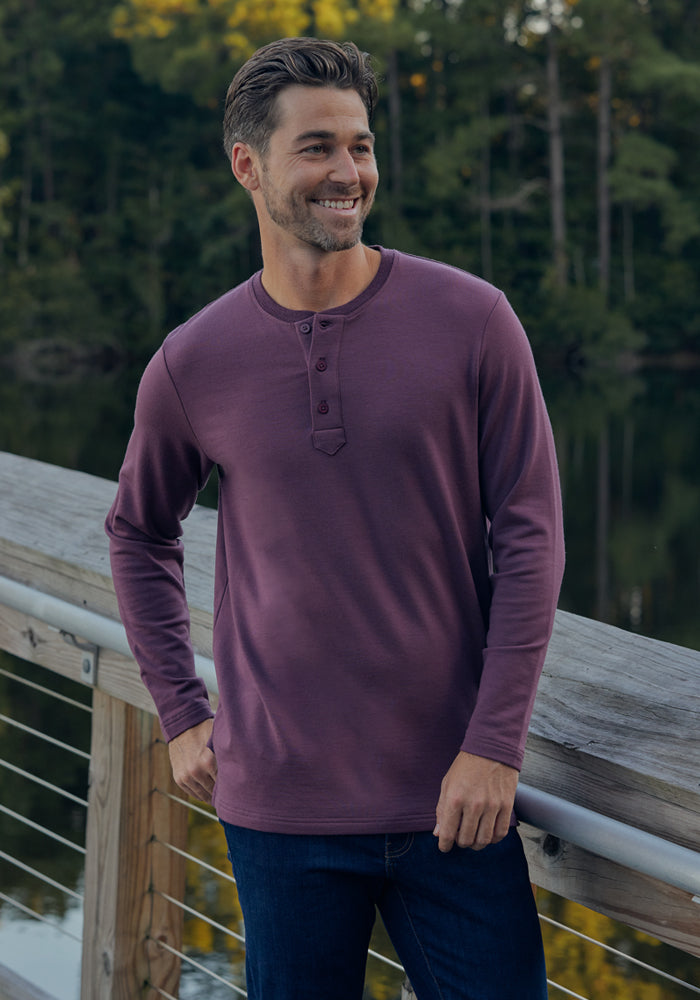 A smiling man with short hair and a beard stands outdoors on a wooden bridge, wearing the Woolx Jackson Henley in black and dark jeans, with trees and water visible in the background.